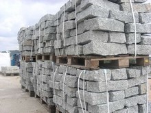 Szare, granitowe bloki ułożone w stosy na drewnianych paletach, zabezpieczone białymi taśmami. Widok z boku, w tle zieleń i fragment niebieskiego pojazdu.